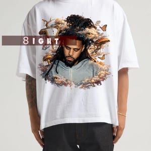 May include: A white short-sleeved t-shirt featuring a graphic print of a person with dreadlocks surrounded by clouds, mushrooms, butterflies, and eyes. The text 8IGHT is printed in white over a dark red rectangular bar across the upper chest area.