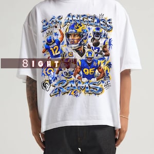 Puede incluir: Una camiseta blanca de manga corta con un estampado gráfico de jugadores de fútbol americano de los Los Angeles Rams con uniformes azules y amarillos. El diseño incluye el texto "LOS ANGELES" y "RAMS" en azul metálico, junto a los números 12, 95 y 17.
