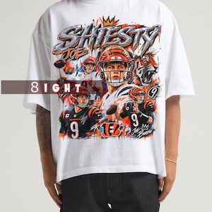 May include: A white short-sleeve t-shirt featuring a graphic print of football player Joe Burrow in a helmet and jersey. The design includes the text SHIESTY, JOE, and 9, along with a tiger illustration, orange and black accents, and a crown icon.