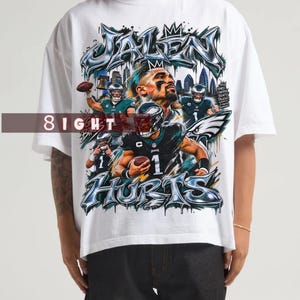 Può includere: Una t-shirt bianca a maniche corte con una stampa grafica di Jalen Hurts in divisa da football. Il design include il testo JALEN HURTS, lo skyline di una città, giocatori di football e la frase HARD WORK BEATS TALENT in toni di blu, nero e argento.