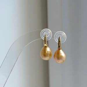 Puede incluir: Un par de pendientes colgantes dorados con perlas doradas en forma de lágrima. Los pendientes tienen un diseño sencillo y elegante con ganchos dorados. Las perlas tienen una superficie lisa y brillante.