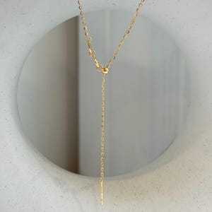 May include: A gold-toned chain necklace featuring a delicate cable link design. The necklace includes an adjustable sliding bead mechanism that allows for length customization, finished with a thin, straight metal bar at the end of the chain.