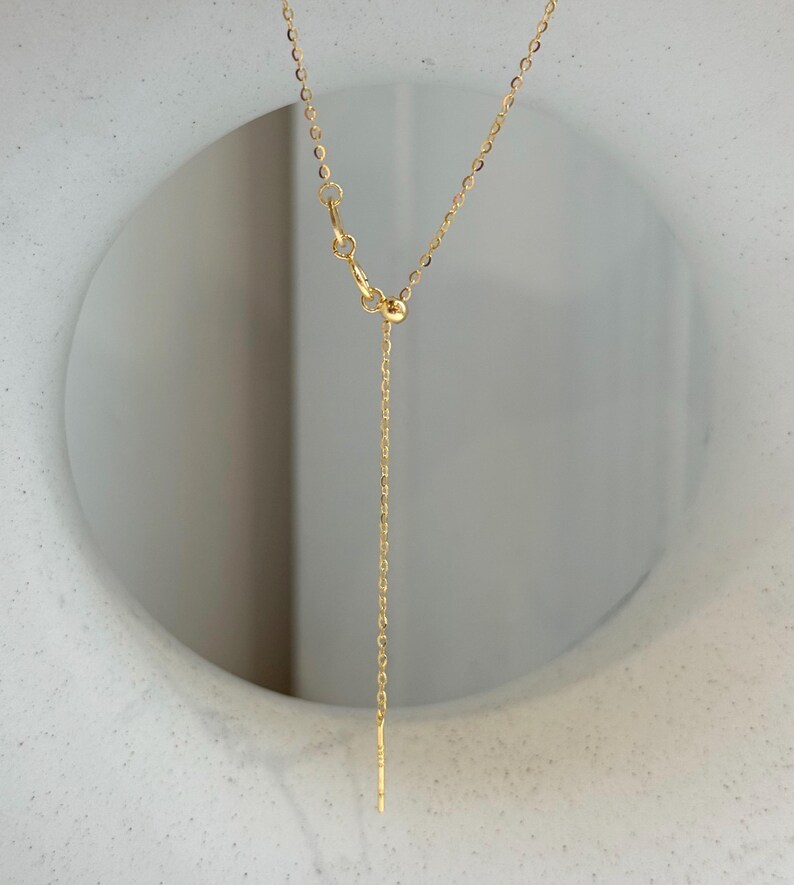 May include: A gold-toned chain necklace featuring a delicate cable link design. The necklace includes an adjustable sliding bead mechanism that allows for length customization, finished with a thin, straight metal bar at the end of the chain.
