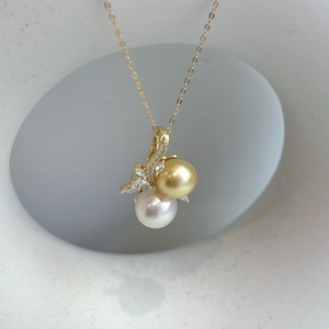 May include: A gold-toned necklace featuring a pendant with two pearls: a white and a golden pearl. The pendant is adorned with small, clear gemstones. The necklace is displayed against a soft, neutral background.