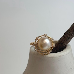 May include: A gold ring featuring a large, round, cream-colored pearl at the center. The pearl is surrounded by a delicate, circular gold frame adorned with small, clear, sparkling stones set at regular intervals around the perimeter.