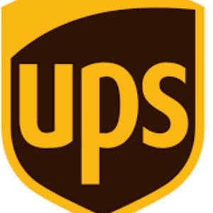 Könnte beinhalten: Das UPS-Logo zeigt ein schildförmiges Design mit einem dunkelbraunen Innenraum und einem goldenen Rand. Die goldenen Buchstaben "ups" sind mittig platziert. Das Design ist einfach und gut erkennbar.