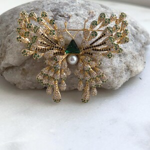 May include: A gold-toned butterfly brooch with intricate detailing. The wings feature green and clear gemstones, with a central emerald-green triangle and a pearl. The brooch is set against a light gray stone.