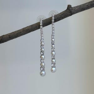 May include: A pair of dangling earrings with a linear design. Each earring features a row of small, sparkling clear stones alternating with round, white pearls. The earrings are suspended from a small, clear stud.