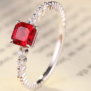 May include: A silver-toned ring featuring a central square-cut vibrant red gemstone held by four prongs. The slender band is accented with a series of small, clear, round-cut stones embedded along the shank on both sides of the main setting.