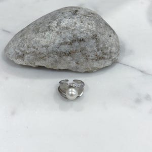 Puede incluir: Un anillo plateado con una perla blanca redonda y grande en el centro. El anillo tiene una banda de pequeños cristales transparentes. El anillo está sobre un fondo blanco con una piedra gris.