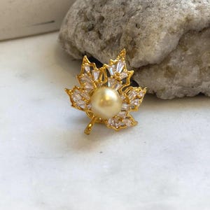 May include: A gold-toned maple leaf brooch featuring a central, round, light-colored pearl. The leaf design is accented with clear, baguette-cut crystals. The brooch is set against a neutral background.
