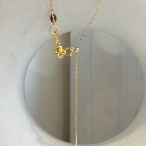 May include: A gold-toned necklace featuring a delicate chain with a spring ring clasp and a small cylindrical pendant at the end. The necklace includes a small circular tag near the clasp and a thin, vertical bar-shaped charm hanging from the chain.