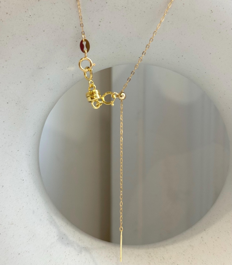 May include: A gold-toned necklace featuring a delicate chain with a spring ring clasp and a small cylindrical pendant at the end. The necklace includes a small circular tag near the clasp and a thin, vertical bar-shaped charm hanging from the chain.