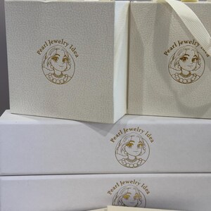 May include: Two cream-colored square gift boxes with ribbon handles and two rectangular white boxes stacked below them, all featuring a gold circular logo of a person wearing a pearl necklace and the text Pearl Jewelry Idea printed on the front.