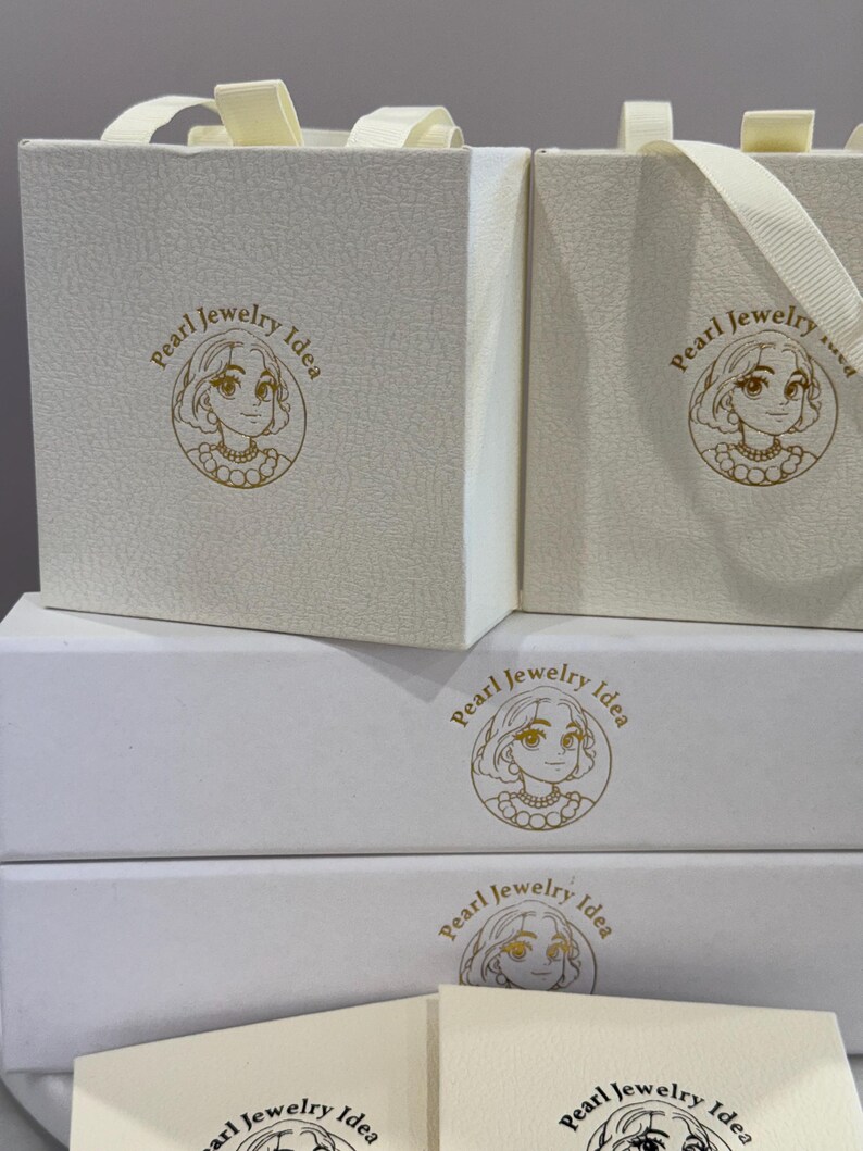 May include: Two cream-colored square gift boxes with ribbon handles and two rectangular white boxes stacked below them, all featuring a gold circular logo of a person wearing a pearl necklace and the text Pearl Jewelry Idea printed on the front.