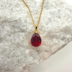 May include: A gold-colored necklace with a teardrop-shaped, faceted, deep red gemstone pendant. The pendant is suspended from a delicate gold chain. The necklace is displayed on a white, textured surface.