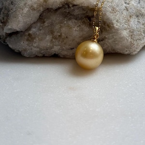 May include: A gold-colored pendant necklace featuring a large, round pearl. The pearl is suspended from a delicate gold chain. The necklace is displayed against a textured, light-colored stone.