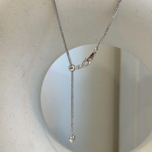 May include: A silver-colored necklace with a delicate chain and a small heart pendant. The necklace features an adjustable length and a secure clasp. The heart pendant is a simple, polished design. The necklace is displayed against a neutral background.