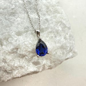 Peut inclure: Un collier en argent avec un pendentif en forme de goutte d'eau, orné d'une pierre précieuse bleu foncé. Le pendentif est serti dans un cadre en métal argenté et est suspendu à une fine chaîne en argent. La pierre précieuse a une surface à facettes.