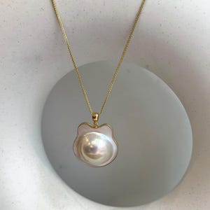 May include: A gold-colored chain necklace with a large, iridescent pearl pendant. The pendant is framed by a gold border and has a unique, irregular shape. The necklace is displayed against a neutral background.