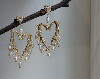 May include: Gold-coloured heart-shaped earrings with dangling white pearls. The earrings feature a textured, organic design and are suspended from small, sparkly, heart-shaped studs. The earrings are displayed against a neutral background.