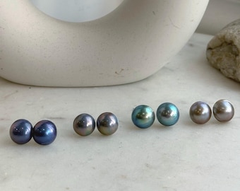 Può includere: Quattro paia di orecchini a lobo rotondi con perle lucenti in varie tonalità di blu scuro, grigio antracite, verde ottanio e bronzo chiaro. Ogni paio è composto da due perle sferiche abbinate con una superficie liscia e riflettente.