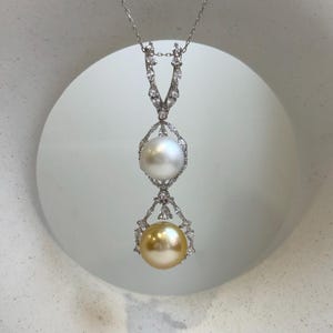 May include: A silver pendant necklace featuring two pearls. The top pearl is white, and the bottom pearl is gold. The pearls are set in ornate, diamond-like settings, suspended from a delicate chain.