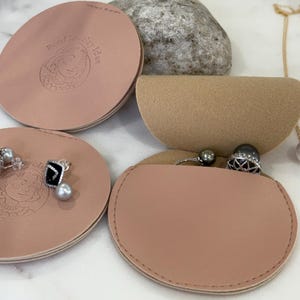 Puede incluir: Organizadores de joyas de cuero rosa con el texto "Pearl Jewelry Idea" y un logotipo. La imagen muestra un par de pendientes con detalles negros y perlas, un anillo con una gema negra y un collar con un colgante de perla.
