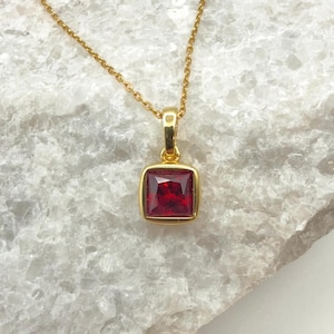 May include: A gold-colored necklace with a square, red gemstone pendant. The pendant is set in a gold frame and hangs from a delicate gold chain. The background is a light-colored, textured surface.