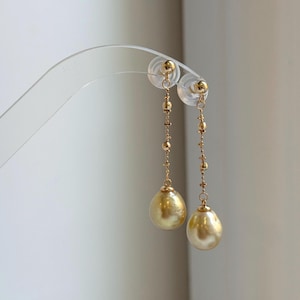 May include: A pair of gold-colored drop earrings. Each earring features a small stud, a delicate chain with small beads, and a large, pear-shaped, golden pearl. The earrings are displayed on a clear earring stand.