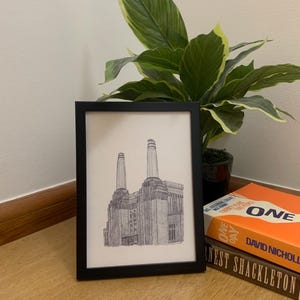 Puede incluir: Lámina artística enmarcada en negro con un dibujo detallado a pluma y tinta de un gran edificio industrial con dos altas chimeneas. La impresión se muestra sobre una mesa redonda, con libros y una planta en maceta en el fondo.
