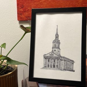 May include: Black framed art print featuring a detailed pen and ink drawing of a church with a tall steeple. The artwork is displayed on a white background and is set against a neutral wall. A potted plant is visible in the foreground.