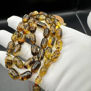 May include: A string of amber prayer beads featuring oval beads in shades of yellow, brown, and black. The beads are strung together with a red string. The background is dark.
