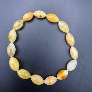 Puede incluir: Un collar compuesto por cuentas ovaladas en tonos amarillos, naranjas y crema. Las cuentas están ensartadas, formando una forma circular sobre un fondo oscuro. Las cuentas tienen un aspecto pulido y brillante. Aproximadamente 40 cm de largo.