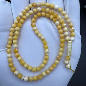 Puede incluir: Un rosario de cuentas amarillas y blancas. Las cuentas están ensartadas, con una borla decorativa en el extremo. Las cuentas son una mezcla de amarillo y blanco, creando un aspecto natural y orgánico.