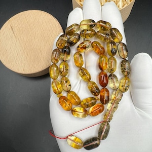 May include: A string of prayer beads featuring translucent, oval-shaped amber beads in varying shades of golden yellow, orange, and brown. The beads are strung on a thin red cord, with a larger, elongated bead at the base and a small tassel.