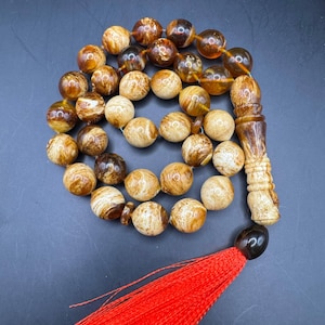 Puede incluir: Un collar de cuentas de oración con cuentas redondas y veteadas en tonos marrón y crema. Las cuentas están ensartadas y terminan con una pieza tallada de color marrón claro y una borla roja. El fondo es gris oscuro.