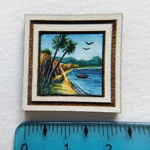 May include: Miniature framed painting of a tropical beach scene. The artwork depicts palm trees, a sandy shore, and a boat on the blue water. The frame is white with a brown inner border. The painting is approximately 1 inch square.