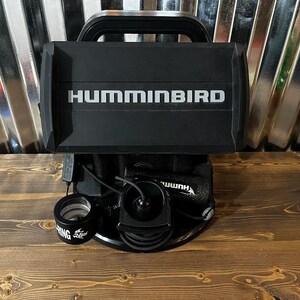 May include: A black Humminbird fish finder mounted on a stand with a black and white Humminbird logo. The fish finder is connected to a black transducer and a black cable.