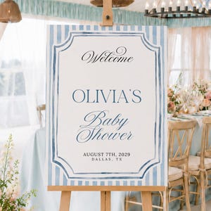K&ouml;nnte beinhalten: Ein rechteckiges Willkommensschild auf einer Holzstaffelei mit einem hellblau-wei&szlig; gestreiften Rand. Der mittlere Text lautet Welcome, OLIVIA'S Baby Shower, AUGUST 7TH, 2029 und DALLAS, TX in einer Mischung aus schwarzen und blauen Schriftarten.