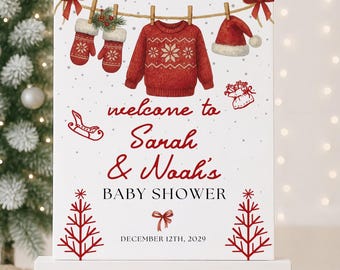 May include: A white sign on an easel with red Christmas-themed illustrations and text. The sign reads "welcome to Sarah & Noah's BABY SHOWER" and "DECEMBER 12TH, 2029". The illustrations include a sweater, mittens, a Santa hat, and Christmas trees.