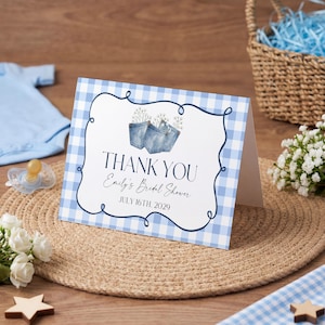 Puede incluir: Una tarjeta de agradecimiento para baby shower con un borde de cuadros azules y blancos y detalles florales. La tarjeta dice "Thank You Emily's Bridal Shower July 16th, 2029." Otros artículos incluyen un body azul, calcetines, un chupete y una cesta de mimbre.