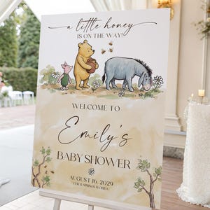 May include: A baby shower welcome sign featuring Winnie the Pooh, Piglet, and Eeyore. The sign reads "a little honey is on the way!" and welcomes guests to Emily's baby shower, scheduled for August 16, 2029.