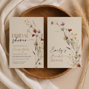A minimalist bridal shower invitation template with a light beige background. On the right side, there is a delicate watercolor arrangement of wildflowers and dried grasses in earthy tones of rust, purple, and cream. The typography is a mix of modern serif and elegant script fonts.