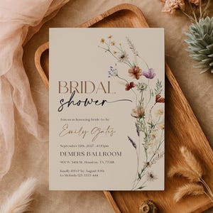 A minimalist bridal shower invitation template with a light beige background. On the right side, there is a delicate watercolor arrangement of wildflowers and dried grasses in earthy tones of rust, purple, and cream. The typography is a mix of modern serif and elegant script fonts.