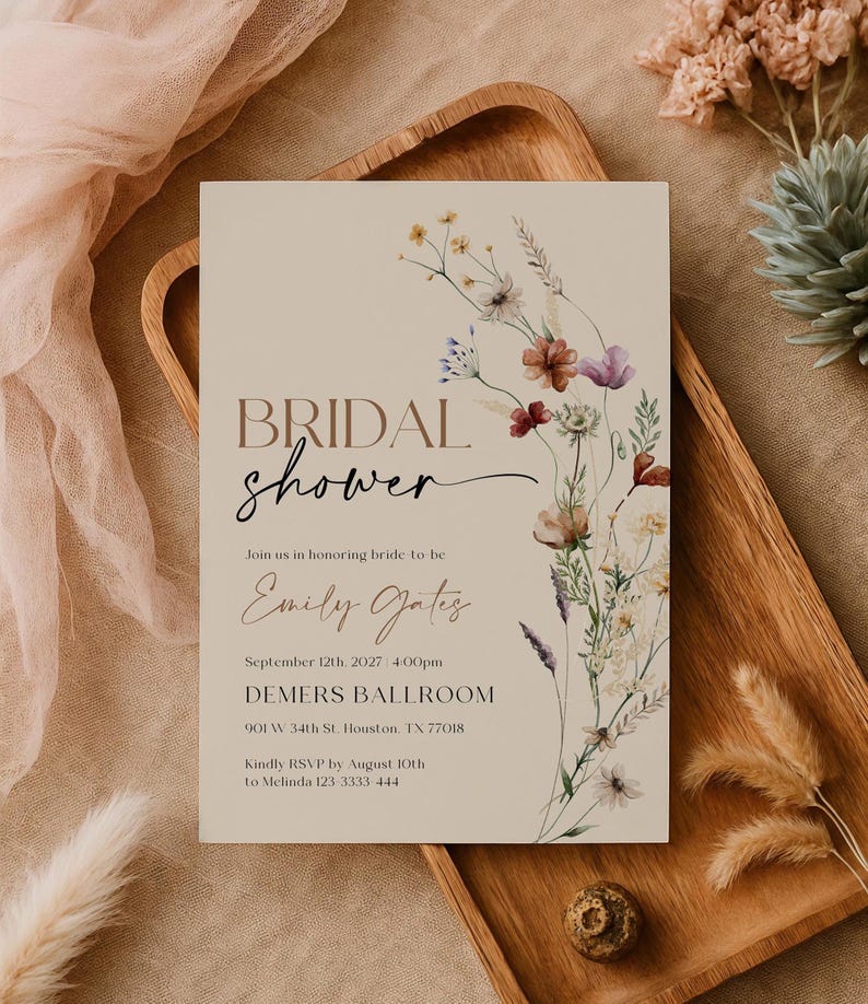 A minimalist bridal shower invitation template with a light beige background. On the right side, there is a delicate watercolor arrangement of wildflowers and dried grasses in earthy tones of rust, purple, and cream. The typography is a mix of modern serif and elegant script fonts.