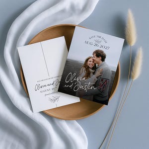 May include: Save the date cards featuring a couple's photo, with the names "Olivia and Justin" in elegant script. The cards are white with a minimalist design, including the date "AUG-26-2027" and a floral accent. The cards are displayed on a wooden tray.
