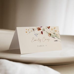 May include: A white place card with a floral design in shades of red, orange, and yellow. The text "Emily Cruz" and "Table 1" is printed in brown.