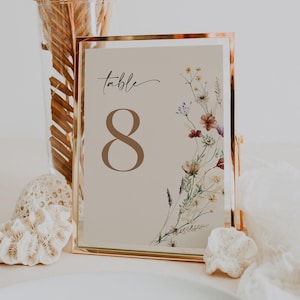 Könnte beinhalten: Eine Tischnummerkarte mit goldenem Rahmen und beigefarbenem Hintergrund. Die Nummer "8" ist in brauner Schrift gedruckt. Die Karte ist mit einer Aquarell-Illustration von Wildblumen in Rosa-, Lila- und Weißtönen verziert.