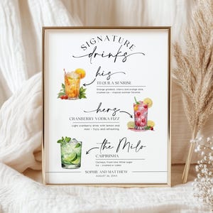 May include: A framed sign with gold trim displays a list of signature drinks. The sign features watercolor illustrations of cocktails: a Tequila Sunrise, a Cranberry Vodka Fizz, and a Caipirinha. The text includes drink ingredients and the names of the couple.